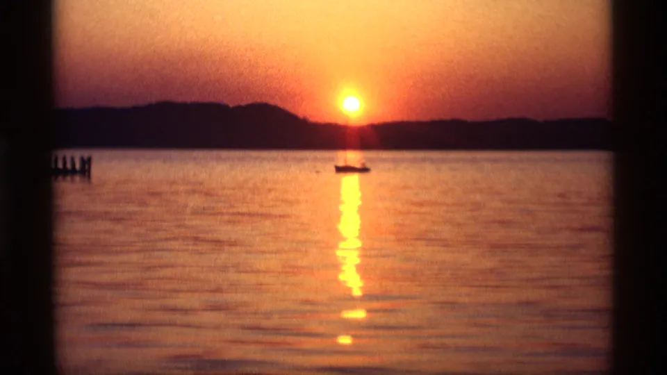 1969 Holland, Michigan home-movie frame of a small silhouetted boat below the setting sun, with lake water and dark shoreline.