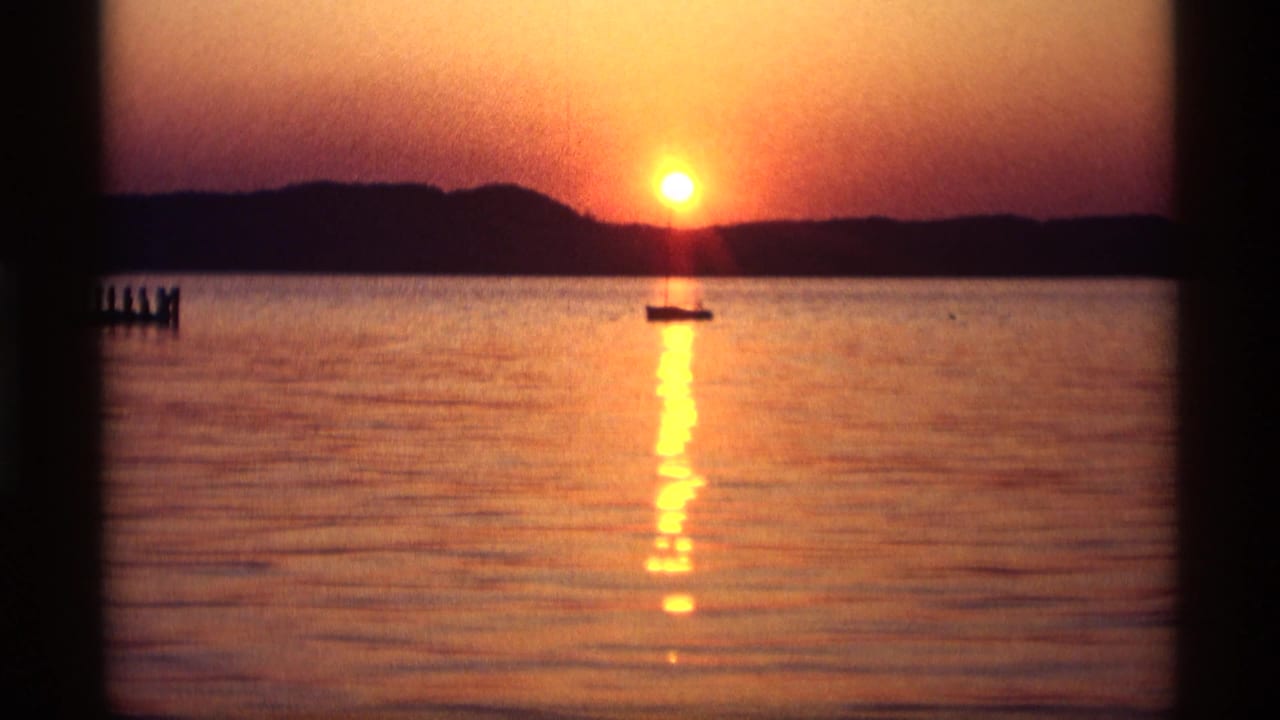 Low sun over water with a silhouetted sailboat and pier at the left edge.
