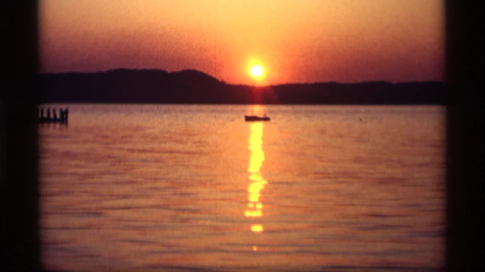 1969 Holland, Michigan 8mm frame with a bright orange sun, rippling reflection on the lake, a silhouetted boat, and dark shoreline.