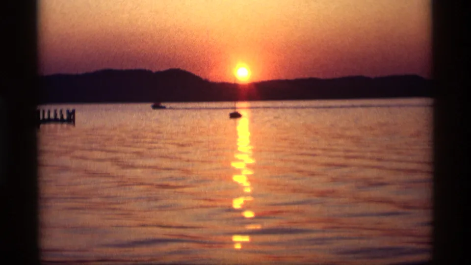 1969 Holland, Michigan 8mm frame showing a warm sunset, two distant boats, shimmering water, and the dark outline of the far shore.