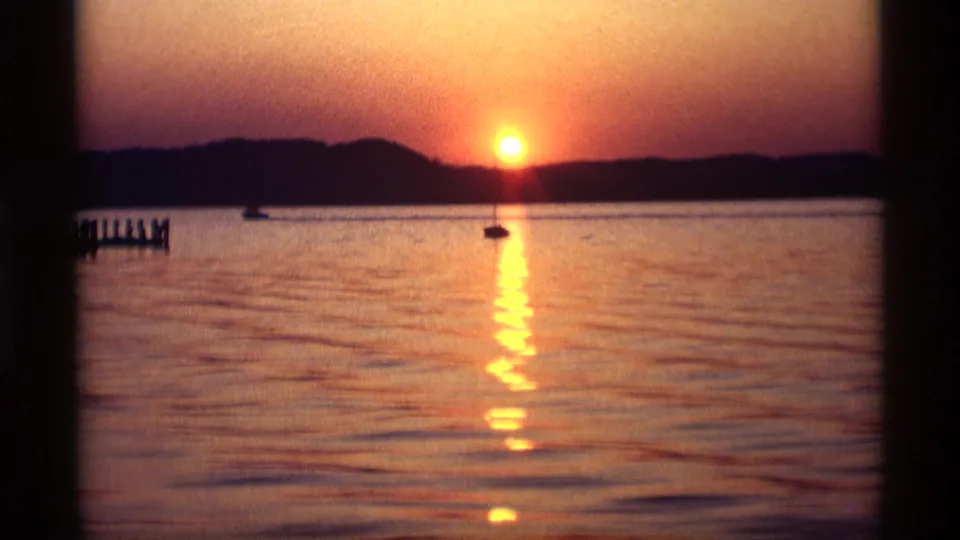 1969 Holland, Michigan 8mm frame showing the sun over the lake, a small silhouetted boat, a pier at left, and warm reflected light on the water.
