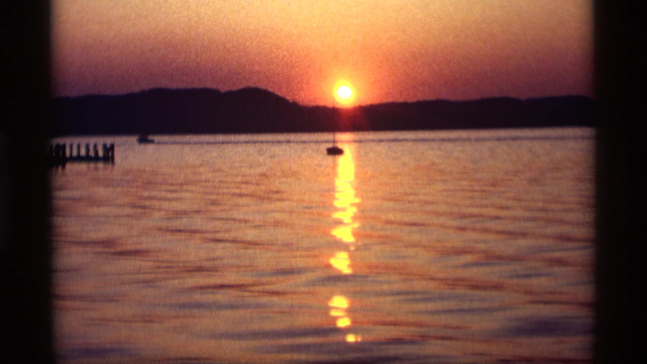 Sunset over calm water with a dock at left and a small distant boat.
