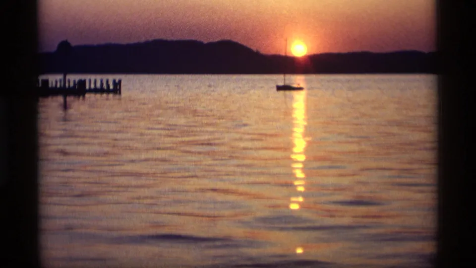 1969 Holland, Michigan 8mm frame showing a bright sun above the shoreline, a sailboat below it, and a dock at the left edge.