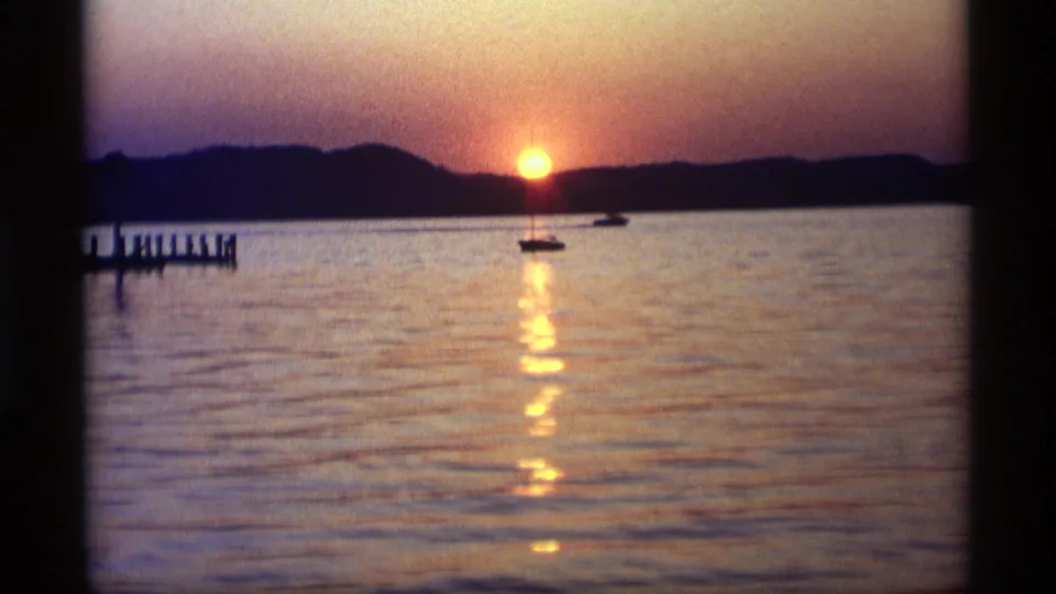 1969 Holland, Michigan lake sunset with two small boats on the water, a dark shoreline, and a pier silhouetted along the left edge.