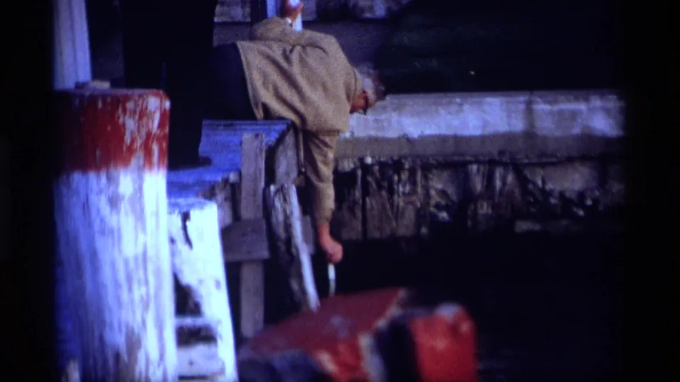 1969 Holland, Michigan 8mm frame showing a man in a light jacket bending over the edge of a pier near painted pilings and dark water.
