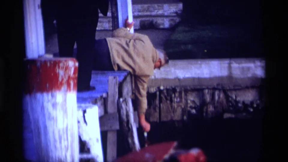 1969 Holland, Michigan pier scene with a man leaning down toward the water, painted pilings in the foreground, and shoreline structures behind him.