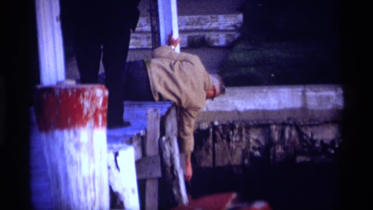 Man in a beige jacket leaning over a weathered dock beside dark water.