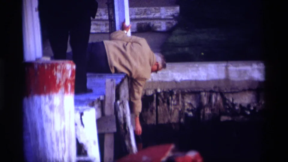 The 1969 Holland, Michigan clip shows the man reaching down from the dock edge, with weathered paint and water visible below.