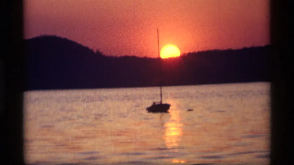 A low orange sun glows over calm water in the Holland, Michigan footage, with a small sailboat silhouetted near the reflection.