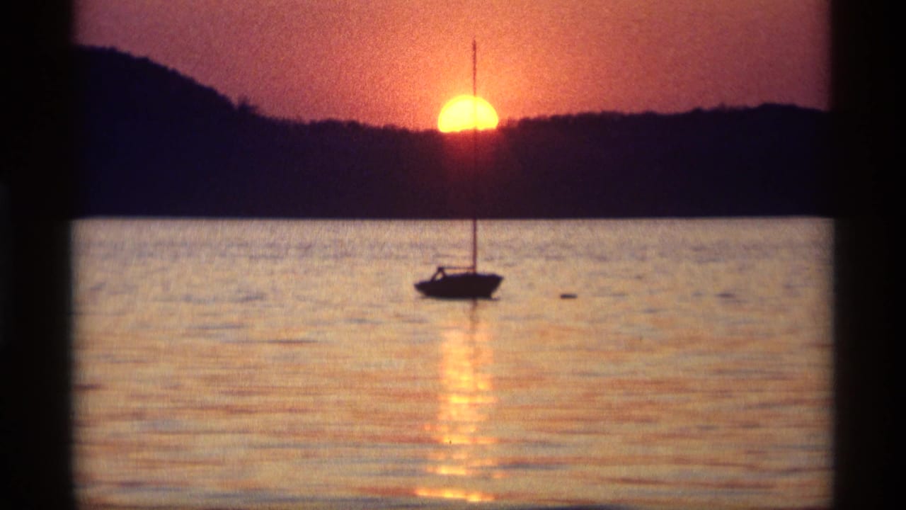 Silhouetted sailboat on calm water beneath a low orange sunset behind dark hills.