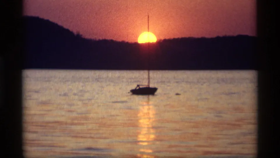 A vintage 8mm view of a silhouetted sailboat on Holland, Michigan water with a gold-orange sun setting behind distant hills.