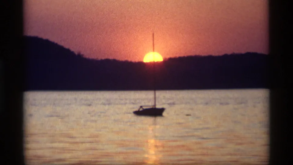 1969 Holland, Michigan home-movie frame with a small sailboat silhouetted below a bright sunset over calm lake water.