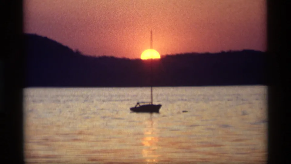Holland, Michigan 8mm sunset frame showing one sailboat on reflective lake water beneath a dark shoreline and orange sky.