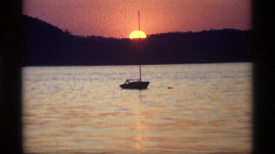1969 Holland, Michigan 8mm frame with a dark sailboat below a glowing sunset and distant tree-lined shore.