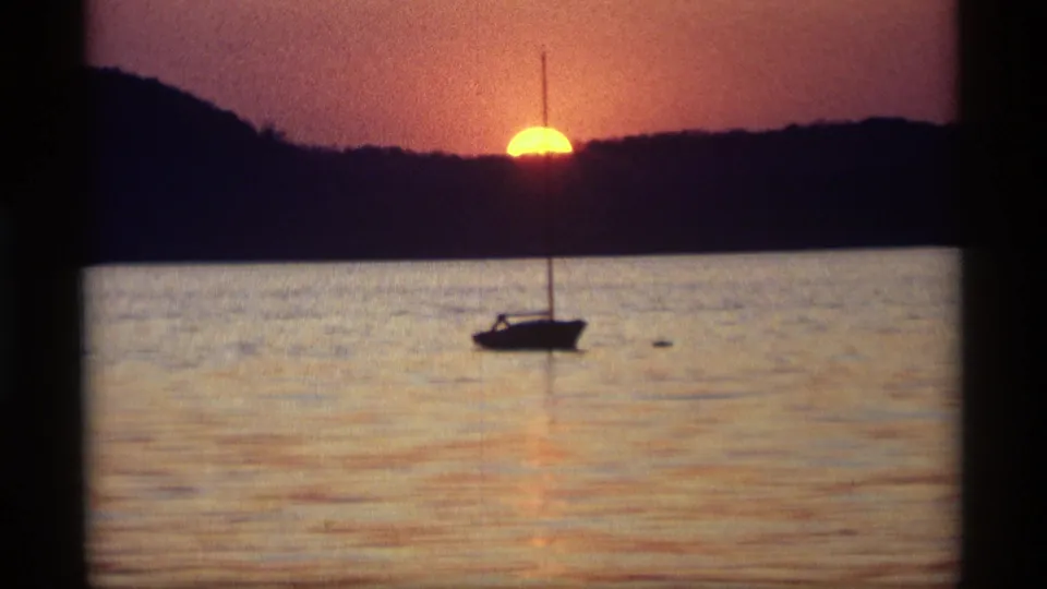 1969 Holland, Michigan home-movie frame showing a sailboat silhouette beneath a low sun over calm water.