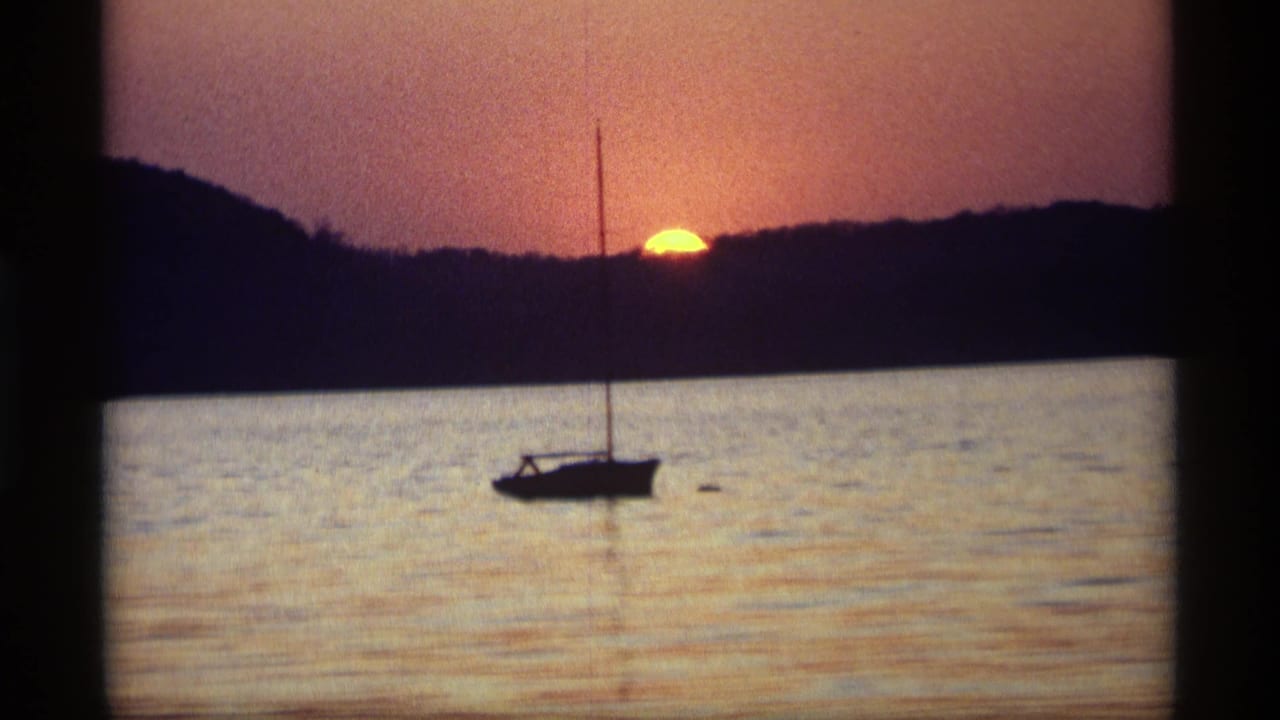 Silhouetted sailboat on calm water below an orange sunset and dark treeline.