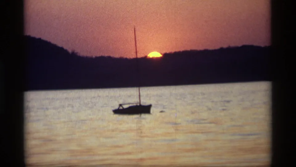 1969 8mm Holland, Michigan sunset view with a dark sailboat and mast on still water beneath a wooded horizon.