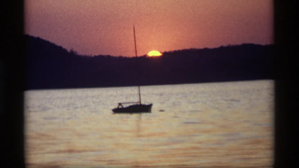 A 1969 Holland, Michigan home-movie frame holds on a silhouetted sailboat as the orange sun nears the treeline over calm water.
