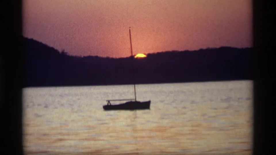 1969 8mm frame from Holland, Michigan showing a small sailboat in silhouette with sunset color over water and a wooded horizon.