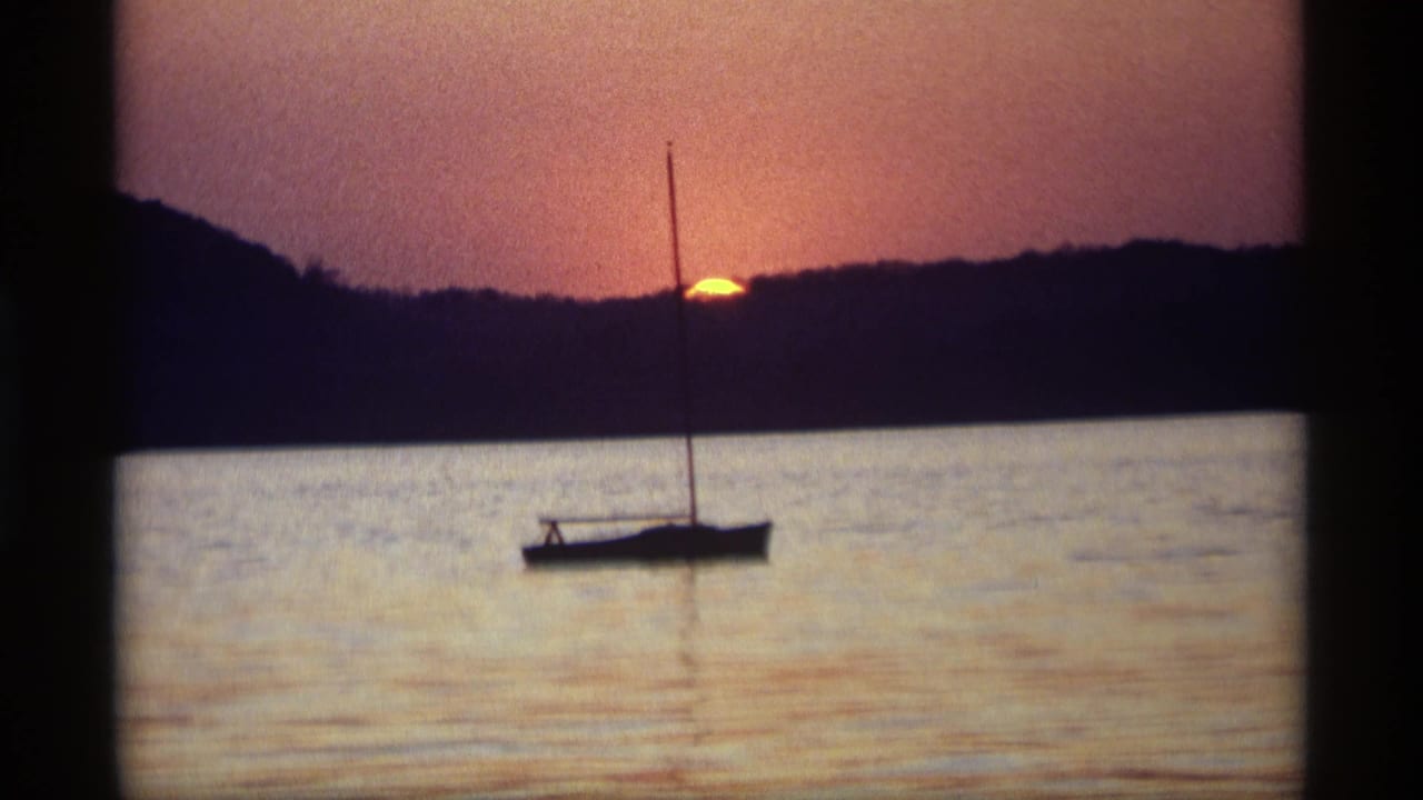 Sailboat silhouette on calm water as the sun sets behind a dark shoreline.
