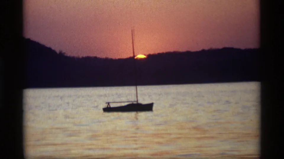 1969 Holland, Michigan home-movie frame with a sailboat in silhouette, calm water, and the sun partly hidden by the far shore.