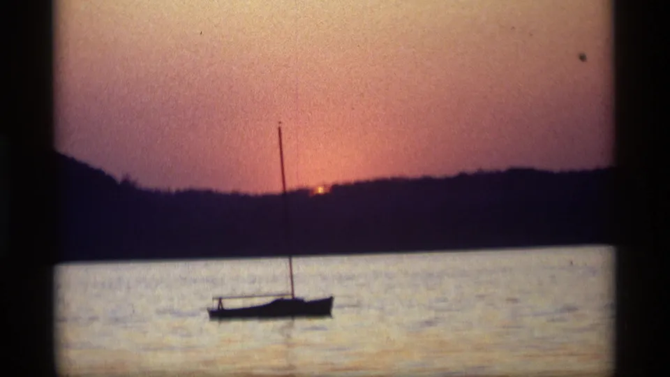 A sailboat sits in silhouette on a calm Holland, Michigan lake as the 1969 sunset glows behind the far shore.