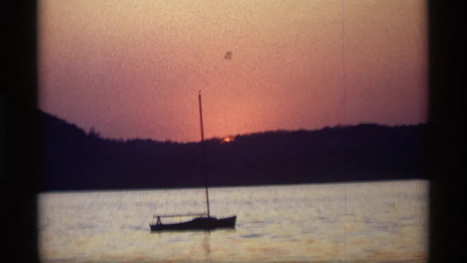 1969 home-movie frame from Holland, Michigan showing a dark sailboat on a lake as the sun meets the distant treeline.