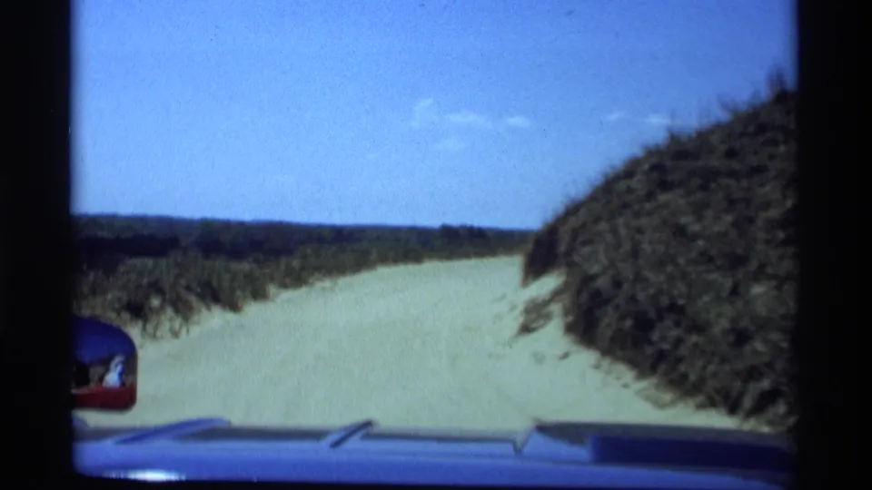 Windshield view of a sandy dune road in Holland, Michigan, with car hood and side mirror visible in the 1969 8mm frame.