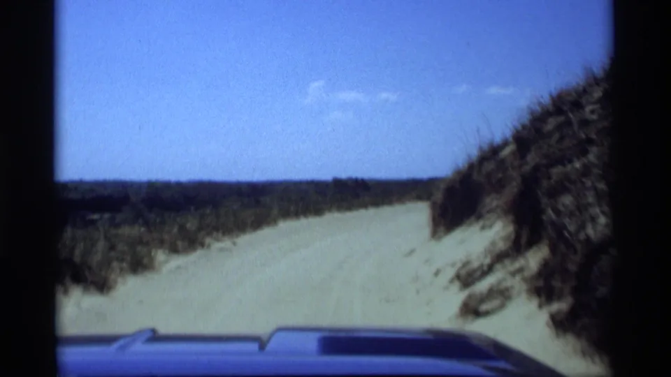 1969 Holland, Michigan home-movie view over a car hood as a sandy road curves through grassy dunes.