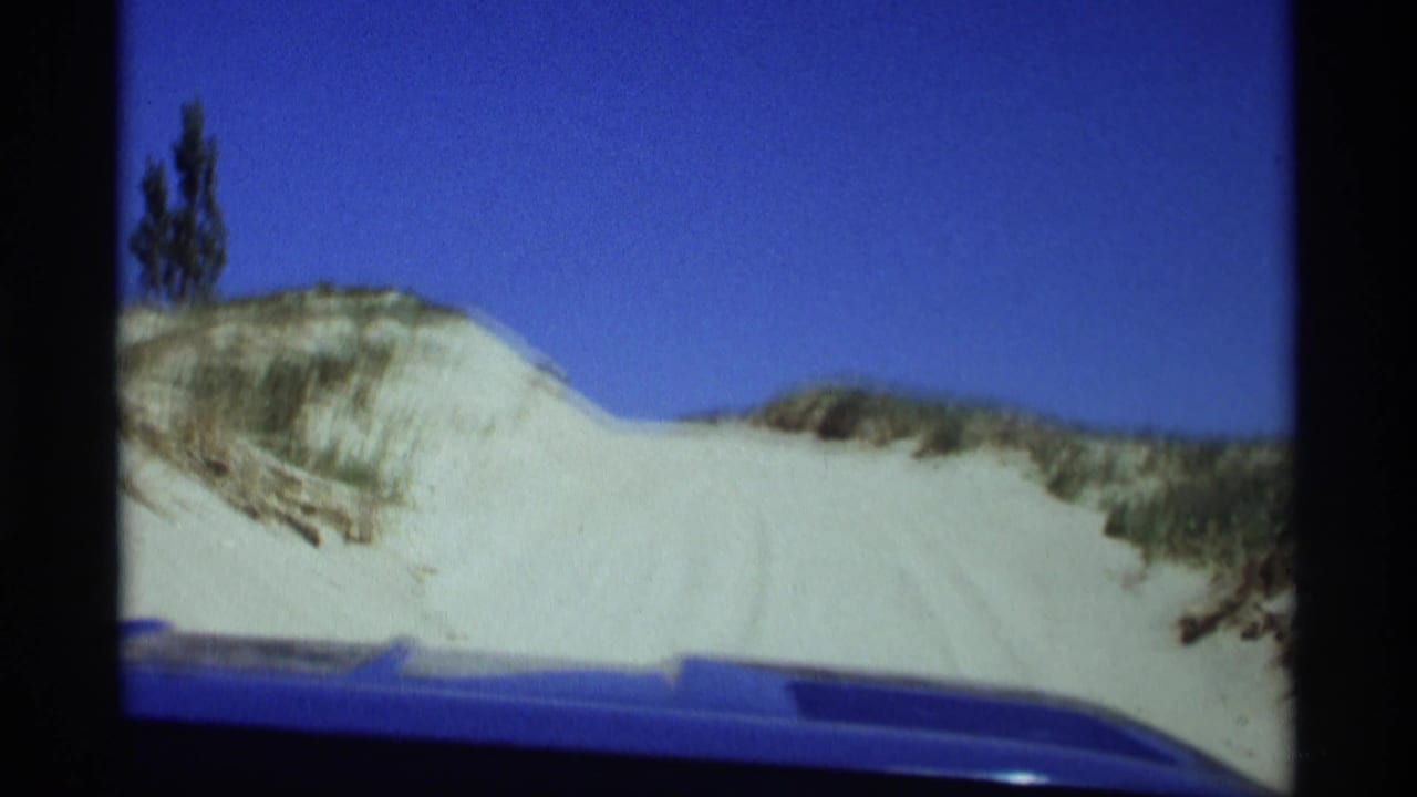 Blue car hood facing pale sand dunes under a clear blue sky.