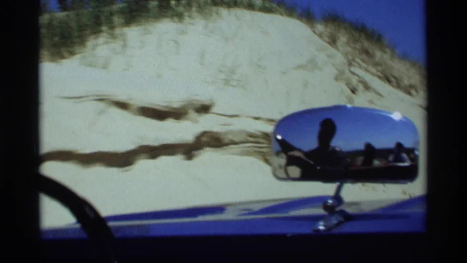 A chrome side mirror on the blue car reflects occupants while the vehicle passes sand dunes in Holland, Michigan.