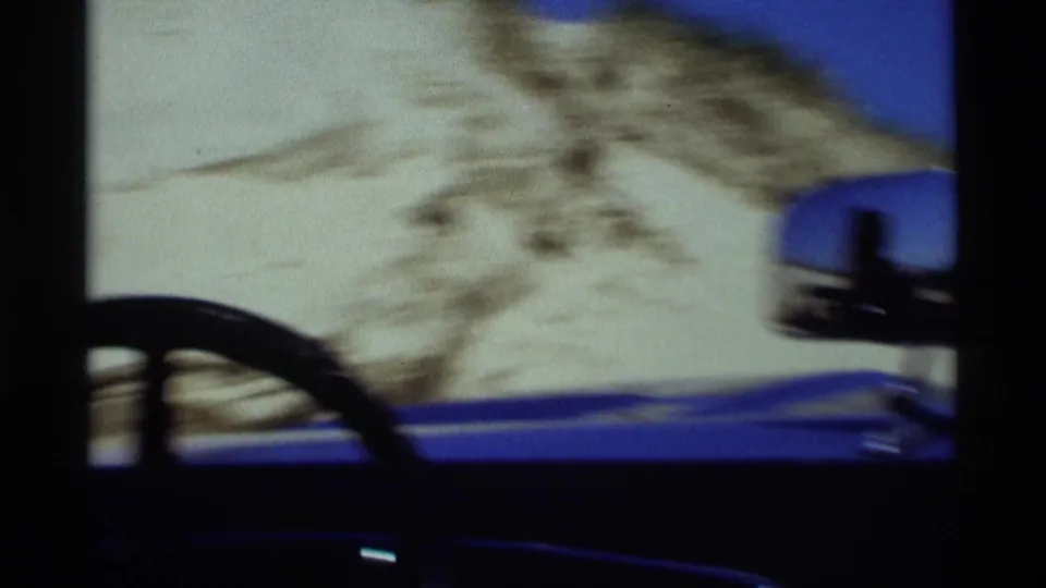 Motion-blurred 8mm view from the blue car as it travels beside sand dunes, with the steering wheel and mirror partly visible.