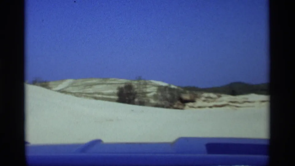 Wide home-movie view over the blue car hood toward pale dunes, sparse vegetation, and distant low hills in Holland, Michigan.