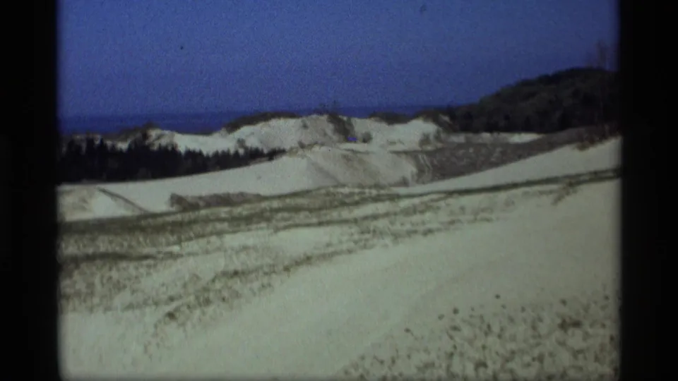 1969 Holland, Michigan 8mm frame showing pale dunes, sparse vegetation, a dark tree line, blue sky, and distant Lake Michigan water.