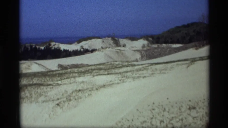 1969 Holland, Michigan 8mm view across rolling sand dunes, scrub vegetation, dark trees, and distant Lake Michigan beneath a clear sky.