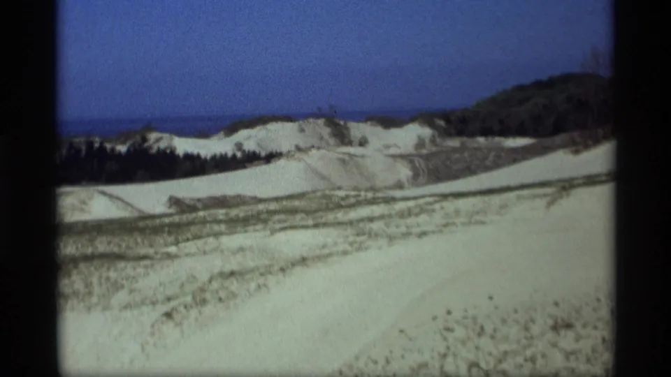 1969 Holland, Michigan home-movie frame of dune ridges and grassy patches leading toward dark trees and distant Lake Michigan water.