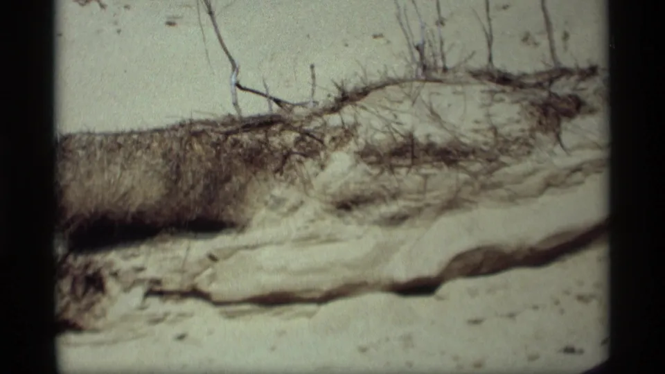 1969 Holland, Michigan 8mm frame showing a closer dune slope with dry brush, exposed sand ledges, grasses, and no visible people.