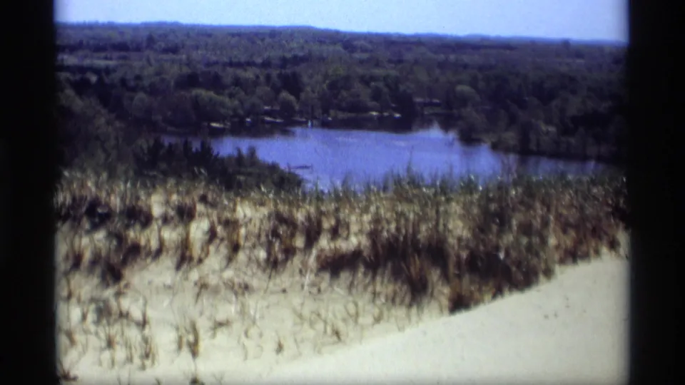 1969 Holland, Michigan 8mm frame with wind-swept dune grass overlooking a calm lake and tree-lined shoreline.