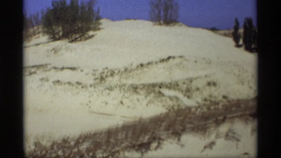 1969 Holland, Michigan frame showing a rounded pale sand dune with sparse brush and grass against a blue sky.