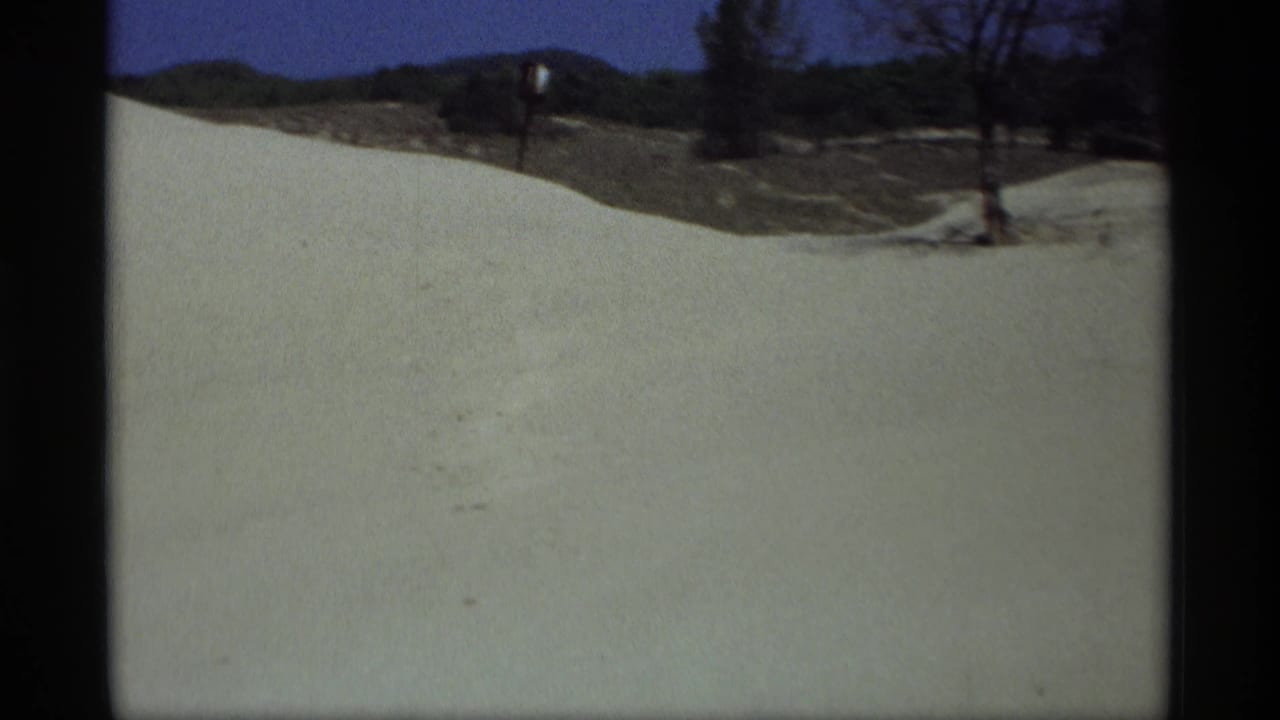 Snow-covered ground beside a dark road edge with bare trees and low hills in the distance.