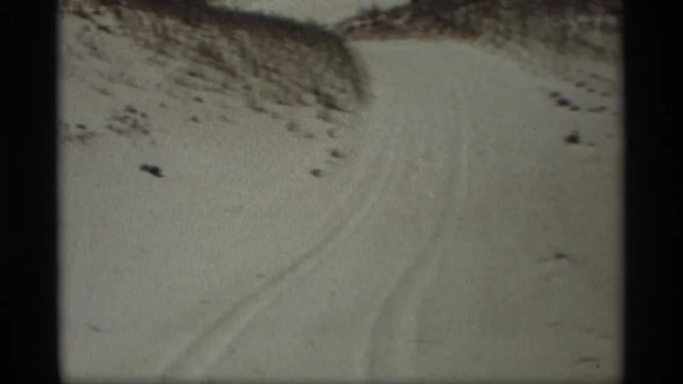 1969 8mm frame from Holland, Michigan showing tire tracks through snow between low sandy or grassy rises.