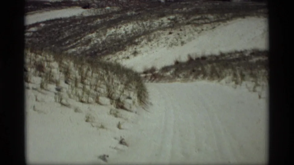 1969 8mm frame from Holland, Michigan showing snow-covered hills, exposed grass, and tracks across the winter landscape.