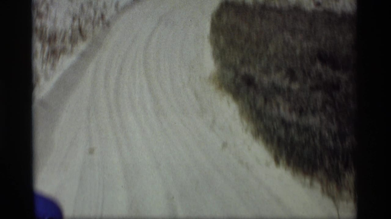 1969 Holland, Michigan 8mm frame showing a pale dirt road bending past roadside brush from a car-travel viewpoint.