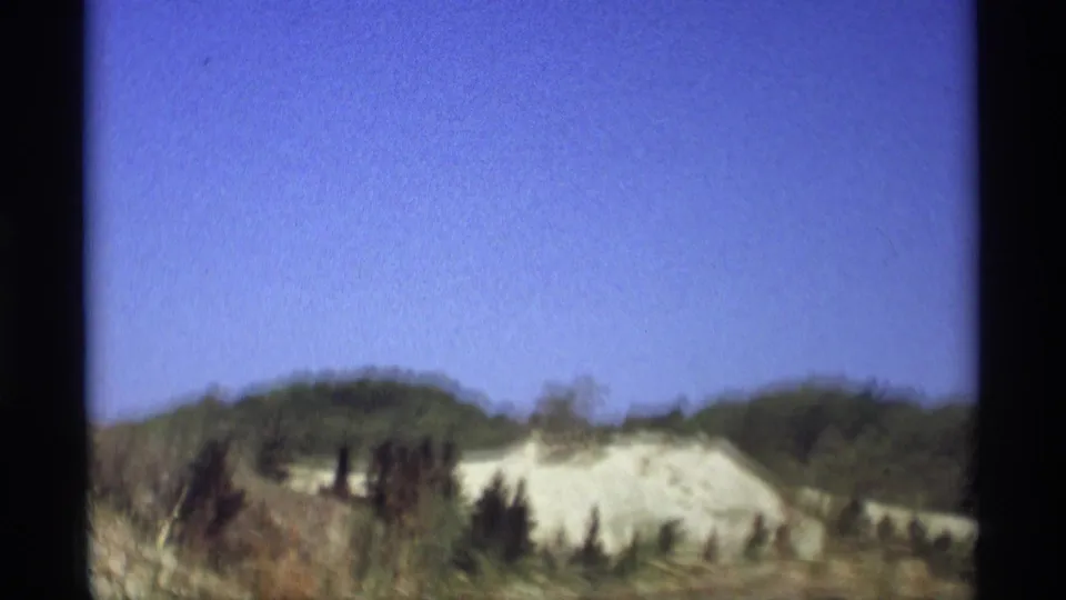 Passenger-window view of low trees, brush, and pale dunes under a clear sky in 1969 Holland, Michigan home-movie footage.