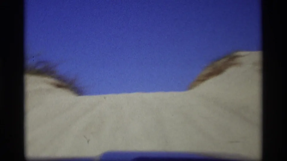 Pale sand dune slopes fill the lower frame beneath a blue sky, with soft 8mm color and slight motion blur.