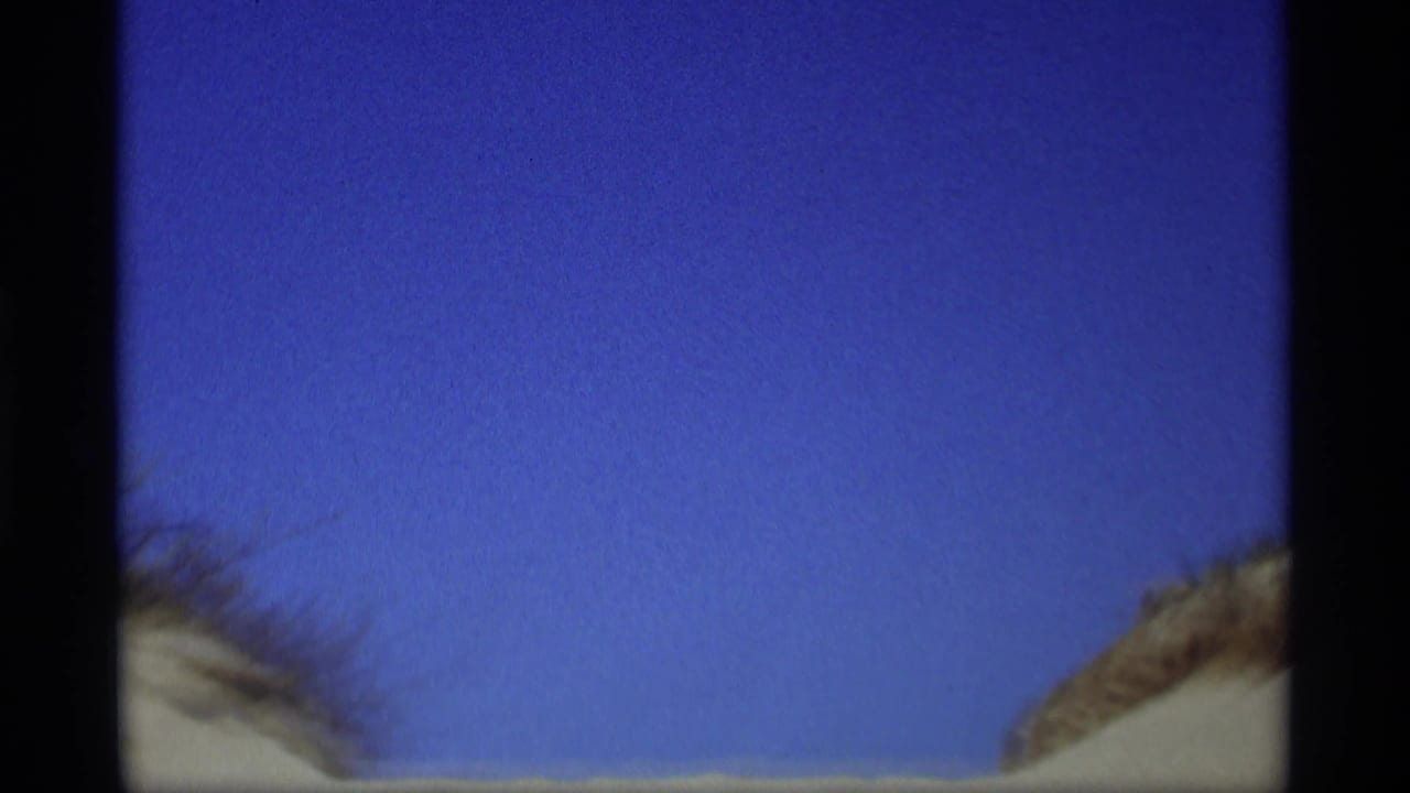 1969 8mm frame from Holland, Michigan showing blue sky above sandy dune edges and heavy gate shadows.