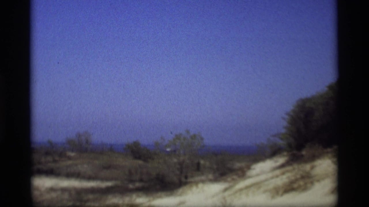 1969 Holland, Michigan 8mm frame showing a sandy dune edge, scrub vegetation, and a broad blue sky.