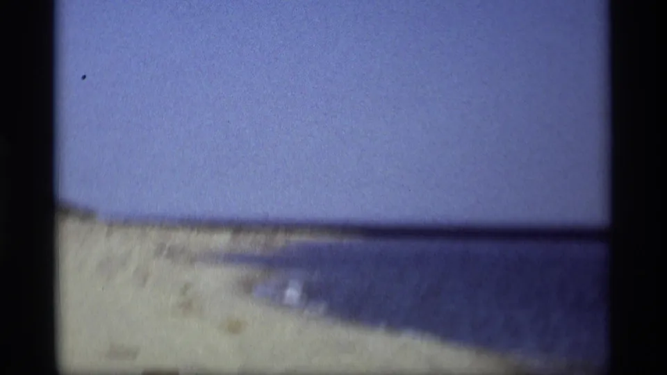 1969 8mm home-movie frame from Holland, Michigan showing a quiet sandy beach and calm water under a pale blue sky.