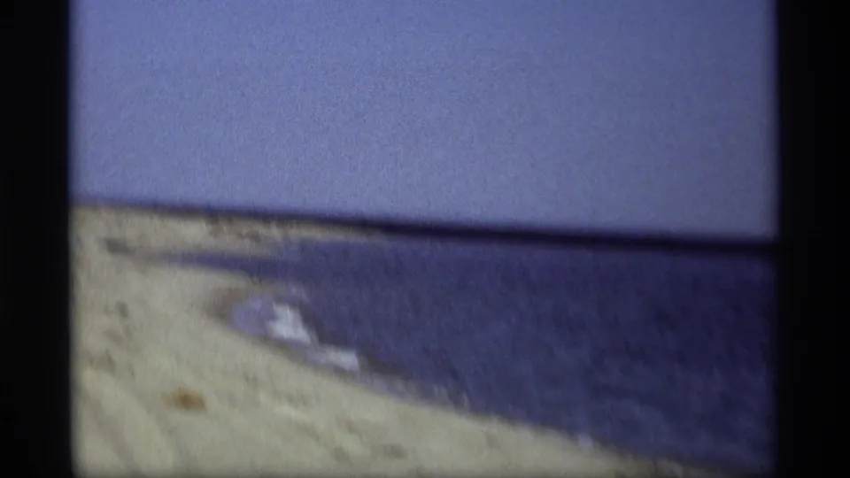 A handheld 1969 Holland, Michigan beach frame shows sand, calm lake water, and a low horizon with visible film texture.