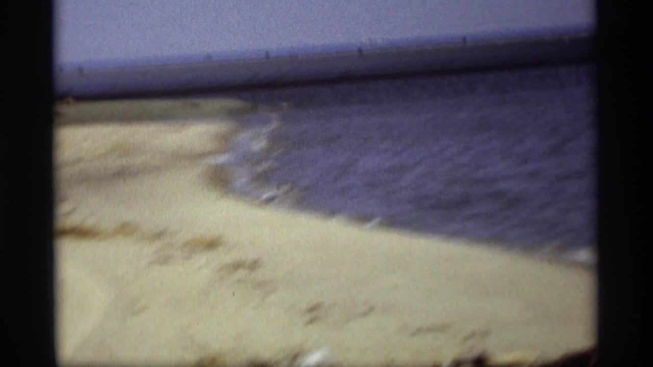 Holland, Michigan lakeshore footage shows pale sand, blue water, and a small shoreline curve in a soft 8mm frame.